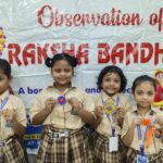 Four smiling schoolgirls in beige uniforms hold colorful handmade crafts in front of a banner